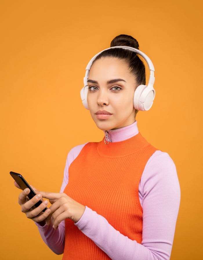 services-04 A young woman listens to music on wireless headphones while using a smartphone against an orange background.